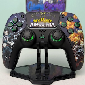 May include: A black wireless video game controller featuring My Hero Academia graphics, with Izuku Midoriya on the left and Katsuki Bakugo on the right. The controller has green backlit buttons and joysticks, and is mounted on a black stand.