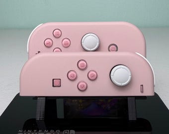 May include: Two pastel pink Nintendo Switch Joy-Con controllers are mounted on a black acrylic display stand. The controllers feature white analogue sticks and pink buttons. The base of the stand is printed with the white Nintendo Switch logo.