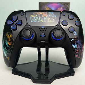 May include: A black PlayStation 5 DualSense controller with a Star Wars themed design. The controller features images of Luke Skywalker, Darth Vader, and the Star Wars logo. The controller is on a black stand.