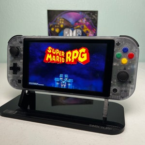 Könnte beinhalten: Eine transparente Nintendo Switch Lite Konsole mit einem schwarzen Ständer. Die Konsole zeigt das Spiel "Super Mario RPG" auf dem Bildschirm an. Die Konsole steht auf einer schwarzen Oberfläche mit dem Schriftzug "Switch Lite".