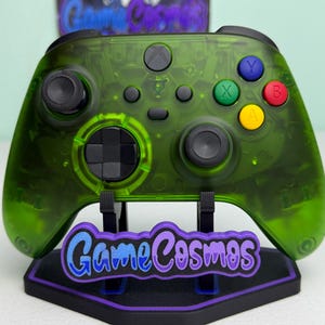 Könnte beinhalten: Ein durchsichtiger, neongrüner Videospiel-Controller mit schwarzen Joysticks, einem schwarzen Steuerkreuz und vier farbigen Aktionstasten mit der Beschriftung X, Y, A und B. Der Controller steht auf einem schwarzen Ständer mit lila GameCosmos-Logo.