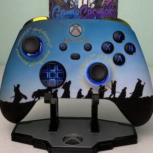 Puede incluir: Mando Xbox personalizado con diseño de El Señor de los Anillos. El mando tiene un fondo degradado azul y blanco con siluetas negras de personajes y detalles dorados. Los botones y joysticks tienen iluminación azul. El mando está sobre un soporte negro.