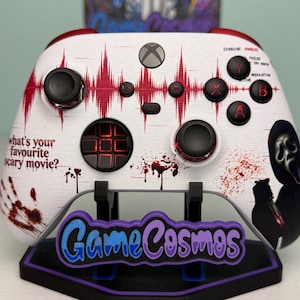 May include: A white wireless game controller featuring a red sound wave graphic, blood splatter patterns, and the text 'what's your favourite scary movie?'. The controller includes a Ghostface character illustration, black buttons, and red accents.