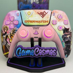May include: A custom pink video game controller featuring a colorful galaxy-themed shell with Overwatch character graphics, the Overwatch logo, and illuminated buttons. The controller is mounted on a black stand with a GameCosmos logo plate.