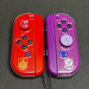 Op de afbeelding: Twee Nintendo Switch-controllers met aangepaste frontplaten. De linker controller is rood met een gouden embleem en een drakenontwerp op de knop. De rechter controller is paars met een zilveren embleem en een drakenontwerp op de knop.