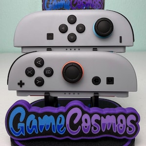 Może przedstawiać: Dwa szare kontrolery do gier z czarnymi przyciskami i joystickami, jeden z niebieskim joystickiem, a drugi z pomarańczowym joystickiem. Kontrolery są ułożone na stojaku z napisem "Game Cosmos" w kolorze niebieskim i fioletowym.