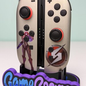 May include: Silver and black game controller with red accents and a purple base. The controller features a Samus Aran graphic and the Game Cosmos logo. The buttons are black, and the joysticks have red rings.