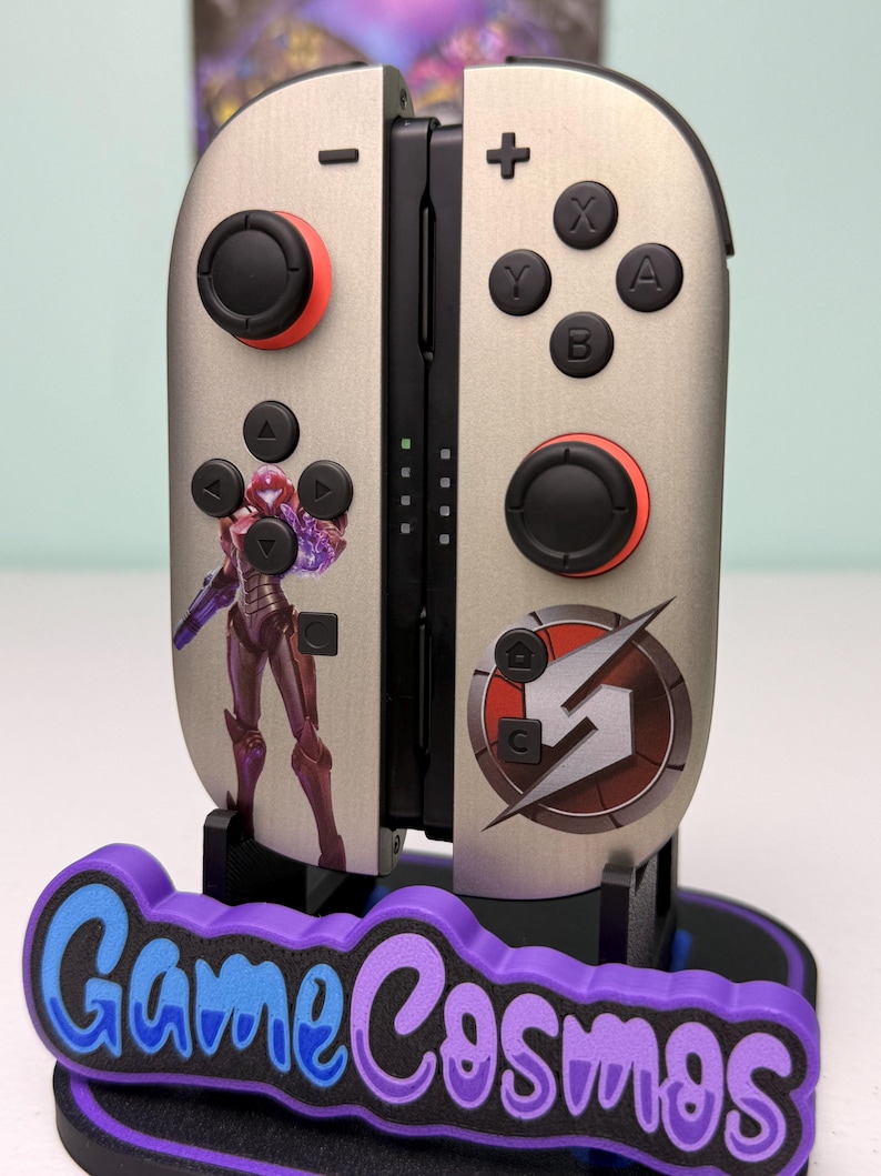 May include: Silver and black game controller with red accents and a purple base. The controller features a Samus Aran graphic and the Game Cosmos logo. The buttons are black, and the joysticks have red rings.