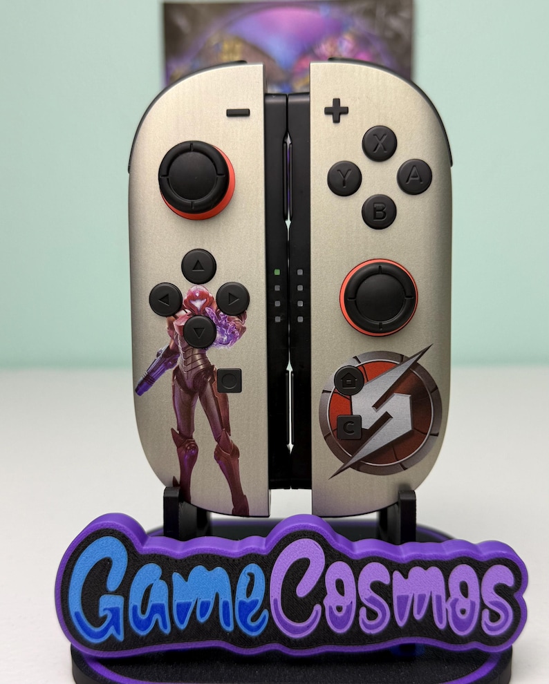 May include: Custom-designed video game controller with a metallic finish. The controller features black buttons and red accents. A graphic of a character is on the left side, and a logo is on the right. The base reads "Game Cosmos" in purple and blue.