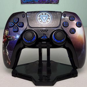 May include: A black PlayStation 5 controller with a custom Iron Man design. The controller has a blue glow around the buttons and is displayed on a black stand. The controller features a galaxy-themed background with Iron Man's arc reactor in the center.