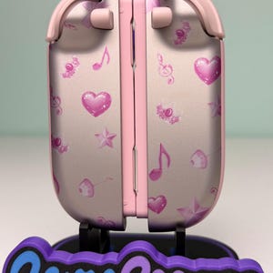 May include: A pink and silver gaming device with a decorative design of hearts, musical notes, and stars. The device is on a stand with the text "Game Cosmos" in blue and purple.