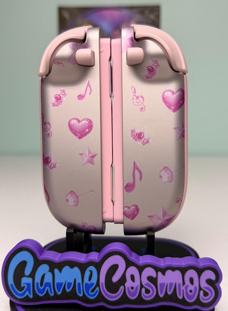 May include: A pink and silver gaming device with a decorative design of hearts, musical notes, and stars. The device is on a stand with the text "Game Cosmos" in blue and purple.