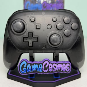 Könnte beinhalten: Ein durchscheinend schwarzer kabelloser Videospiel-Controller mit zwei Analogsticks, einem Steuerkreuz und Tasten mit der Beschriftung X, Y, A und B. Der Controller steht auf einem schwarzen Ständer mit einem lila umrandeten GameCosmos-Logo.