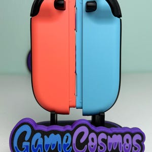 Pu&ograve; includere: Un supporto per controller per console di gioco, con un design del controller corallo e azzurro. Il supporto ha una base nera con il testo "Game Cosmos" in blu e viola.
