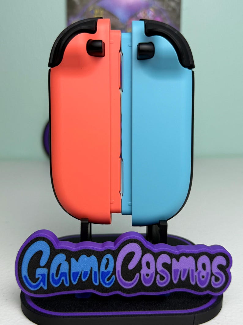 Pu&ograve; includere: Un supporto per controller per console di gioco, con un design del controller corallo e azzurro. Il supporto ha una base nera con il testo "Game Cosmos" in blu e viola.