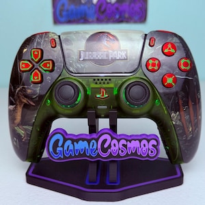 Pode incluir: Um controle de videogame personalizado com o logotipo do Jurassic Park no touchpad, uma carcaça verde escura translúcida com gráficos de dinossauros e botões vermelhos brilhantes com símbolos verdes. O controle repousa sobre um suporte preto com o texto GameCosmos.