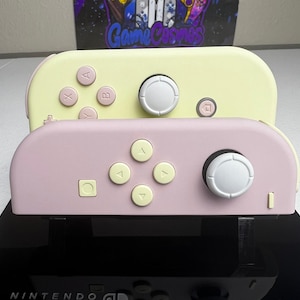May include: Two Nintendo Switch Joy-Con controllers in pastel colors. The top controller is pale yellow with light pink buttons and a white joystick. The bottom controller is pale pink with light yellow buttons and a white joystick.
