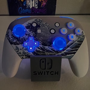 Puede incluir: Un mando blanco de Nintendo Switch con un dise&ntilde;o de olas azul y gris. El mando tiene luces LED azules en los botones y el pad direccional. El mando est&aacute; colocado en un soporte gris con el logotipo de Nintendo Switch y la palabra "SWITCH" impresa en &eacute;l.