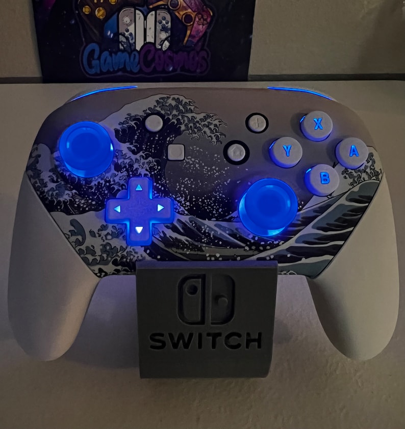 Puede incluir: Un mando blanco de Nintendo Switch con un dise&ntilde;o de olas azul y gris. El mando tiene luces LED azules en los botones y el pad direccional. El mando est&aacute; colocado en un soporte gris con el logotipo de Nintendo Switch y la palabra "SWITCH" impresa en &eacute;l.