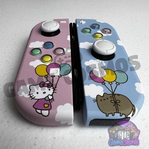 May include: A pair of game controllers featuring a pink left side with Hello Kitty holding balloons and a blue right side with Pusheen holding balloons. Both controllers have white clouds, white joysticks, and colorful buttons.