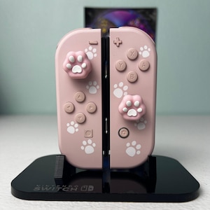 May include: A pair of light pink Nintendo Switch Joy-Con controllers featuring white paw print decals and matching pink and white cat paw thumbstick covers. The controllers are mounted on a black Nintendo Switch branded display stand.