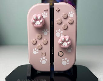 May include: A pair of light pink Nintendo Switch Joy-Con controllers featuring white paw print decals and matching pink and white cat paw thumbstick covers. The controllers are mounted on a black Nintendo Switch branded display stand.