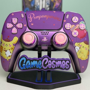 May include: A purple wireless video game controller featuring Pompompurin character graphics, pink joystick caps, and the text Pompompurin printed on the center. The controller is displayed on a black stand with the text GameCosmos in blue and purple.