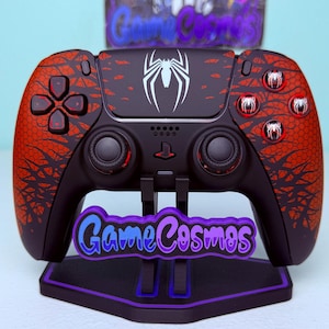 May include: A black wireless video game controller featuring a red and black web-like pattern on the grips, a white spider emblem on the touchpad, and custom buttons with spider icons. The controller rests on a black stand labelled GameCosmos.