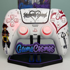 May include: A white wireless video game controller featuring Kingdom Hearts graphics, including a character illustration, a keyblade, and heart symbols. The controller has red glowing analog sticks and buttons, and it rests on a black stand with the text GameCosmos.