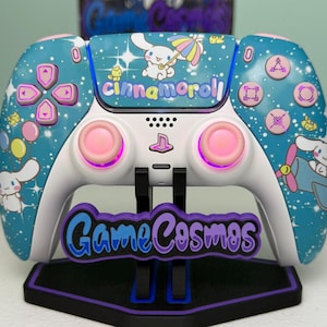 May include: A white and teal wireless video game controller featuring Cinnamoroll character graphics, colorful balloons, and stars. The controller has pink analog sticks, light-up buttons, and the text Cinnamoroll on the touchpad, resting on a black stand.