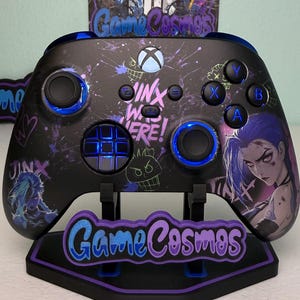 May include: Black Xbox controller with blue detailing and a custom Jinx design. The controller features the text "Jinx Wuz Here!" and is displayed on a stand with the "Game Cosmos" logo.