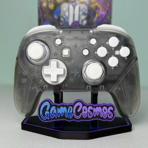 May include: A translucent gray wireless video game controller with white buttons and joysticks, mounted on a black display stand featuring a purple and blue logo that reads GameCosmos in a stylized script font.