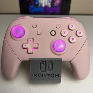 May include: A pink Nintendo Switch controller with a grey stand. The controller has pink buttons and a purple light around the buttons. The stand has the Nintendo Switch logo and the word "SWITCH" printed on it.