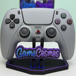 Könnte beinhalten: Ein grauer Videospiel-Controller mit schwarzen Knöpfen und einem Ständer. Der Ständer trägt den Text "Game Cosmos" in Blau und Lila. Der Controller hat das PlayStation-Logo.
