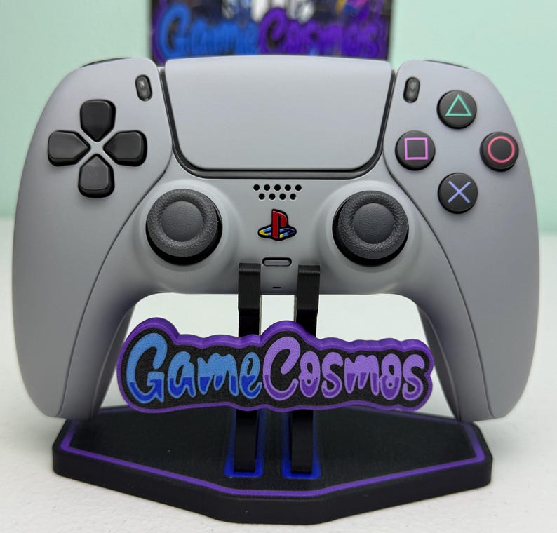 Puede incluir: Un mando de videojuegos gris con botones negros y un soporte. El soporte tiene el texto "Game Cosmos" en azul y morado. El mando tiene el logotipo de PlayStation.