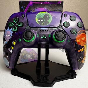 Puede incluir: Un mando de PlayStation 5 morado y negro con un diseño personalizado que presenta personajes de la serie animada Rick y Morty. El mando está en un soporte negro con el logotipo de PlayStation.
