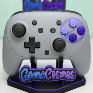 Könnte beinhalten: Ein hellgrauer kabelloser Gamecontroller mit schwarzen Joysticks, einem schwarzen Steuerkreuz und violetten Aktionstasten mit der Beschriftung X, Y, A und B. Der Controller steht auf einem schwarzen Ständer mit violettem GameCosmos-Logo.