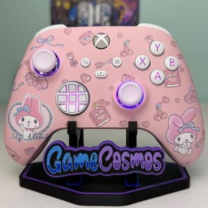 Könnte beinhalten: Rosa Xbox-Controller mit My Melody-Grafiken und weißen Knöpfen. Der Controller hat ein individuelles Design mit verschiedenen Symbolen und dem Text "My Melody". Er wird auf einem schwarzen Ständer mit dem Text "Game Cosmos" präsentiert.