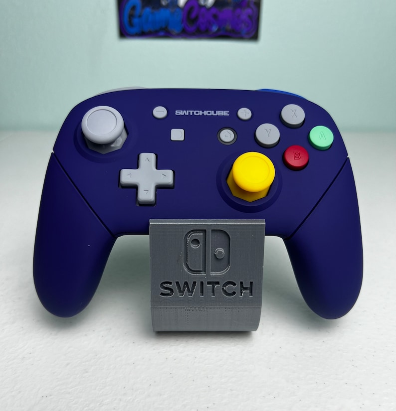 May include: A blue and gray Nintendo Switch controller with a yellow button and the text "SWITCHCUBE" on the front. The controller is sitting on a gray stand with the Nintendo Switch logo and the word "SWITCH" printed on it.