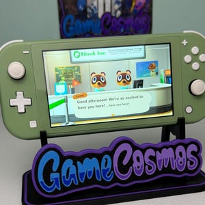 Op de afbeelding: Olijfgroene Nintendo Switch Lite-console met een game-scène met tekst van "Nook Inc." en het personage Timmy. De console staat op een zwarte standaard met de tekst "Game Cosmos" in blauw en paars.