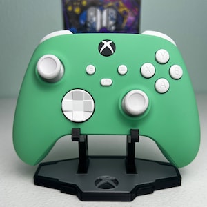 May include: A mint green Xbox controller with white buttons and a white d-pad. The controller is on a black stand with the Xbox logo.