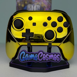 May include: A gold chrome video game controller with black buttons and joysticks, mounted on a black display stand featuring a purple and blue logo plate that reads GameCosmos in a stylized font, centered against a light background.