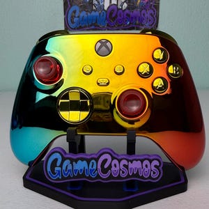 May include: A rainbow-colored Xbox controller with red thumbsticks and gold buttons. The controller is displayed on a stand with the text "Game Cosmos" in purple and blue.