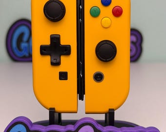 May include: A pair of yellow video game controllers attached to a black central mount. The left controller features a black directional pad and joystick, while the right has a black joystick, four coloured action buttons, and a plus sign button.