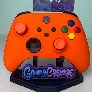 May include: Orange Xbox controller with black accents, resting on a black stand with the text "Game Cosmos" in blue and purple. The controller has colored buttons and a black directional pad.
