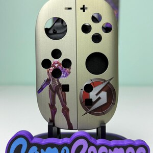 May include: A metallic silver game controller skin with a purple and red graphic of a video game character. The skin has black button holes and a purple and blue "Game Cosmos" logo at the base.