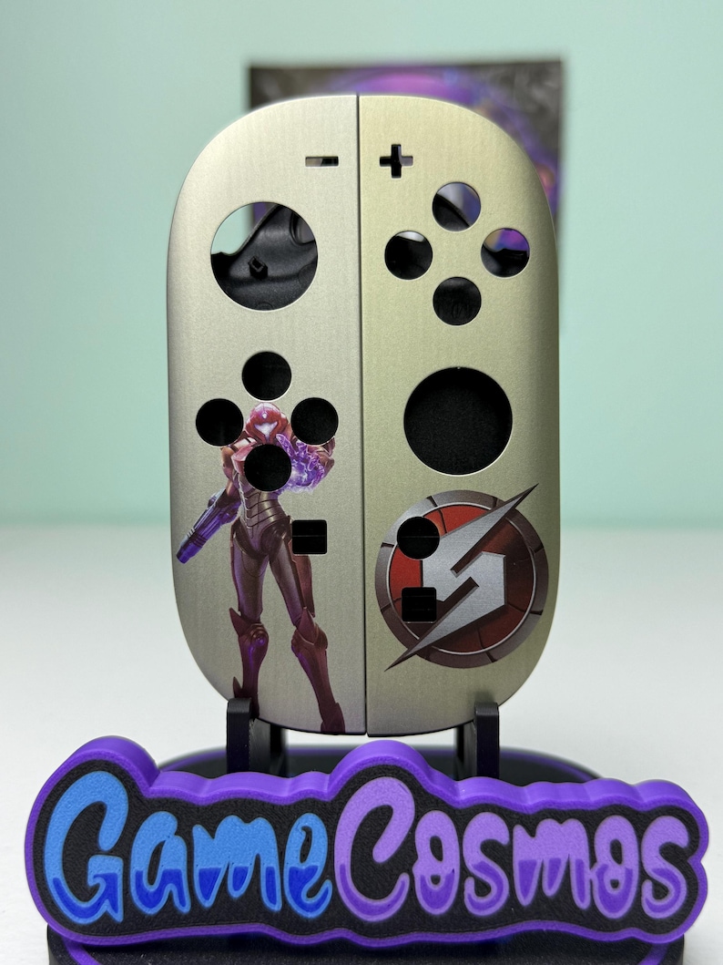 May include: A metallic silver game controller skin with a purple and red graphic of a video game character. The skin has black button holes and a purple and blue "Game Cosmos" logo at the base.