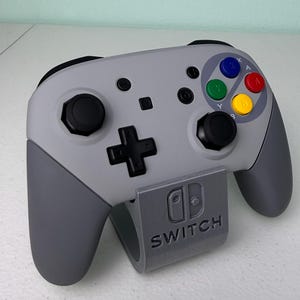 Puede incluir: Un mando gris de Nintendo Switch con una placa frontal de estilo Super Nintendo gris y blanco. El mando est&aacute; colocado en un soporte gris con la palabra "SWITCH" impresa en &eacute;l.