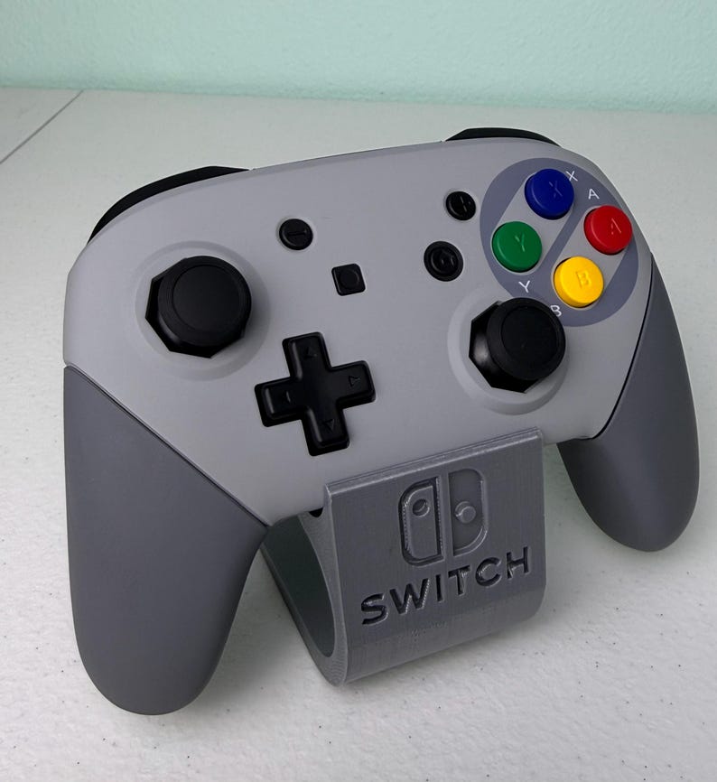 Puede incluir: Un mando gris de Nintendo Switch con una placa frontal de estilo Super Nintendo gris y blanco. El mando est&aacute; colocado en un soporte gris con la palabra "SWITCH" impresa en &eacute;l.