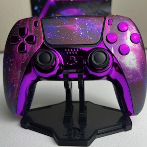 Puede incluir: Un mando de PlayStation 5 morado y negro con un diseño de galaxia en la parte posterior. El mando está en un soporte negro.